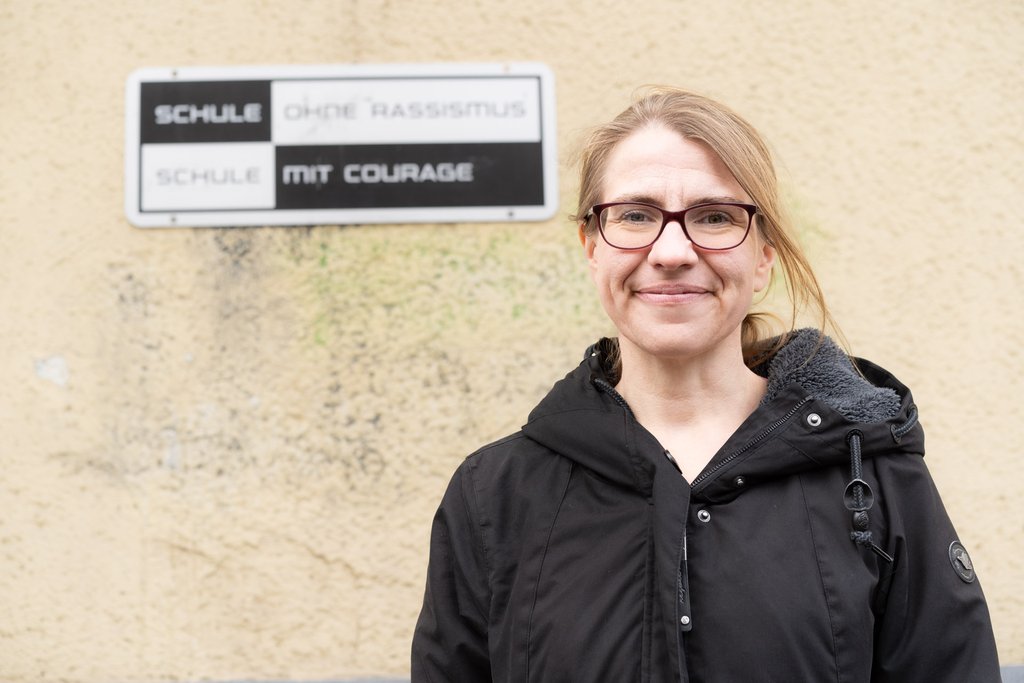 Franziska Brychcy steht vor einer Hauswand an der ein Schild mit "Schule ohne Rassismus Schule mit Courage" hängt.